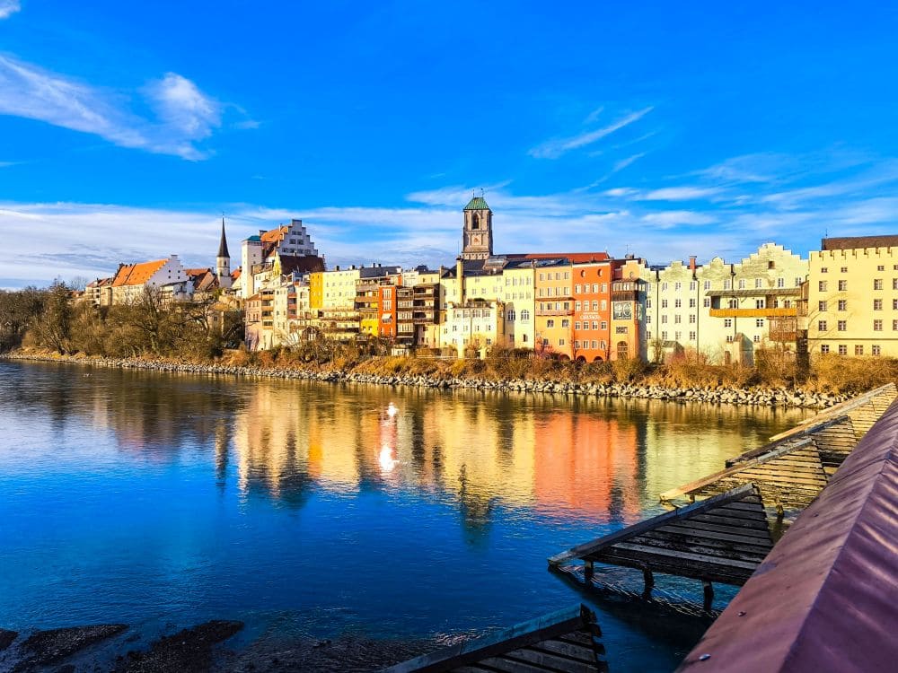 Eine lebendige Flusslandschaft in Wasserburg. Bunte Gebäude spiegeln sich unter einem klaren blauen Himmel im Wasser. Ein Holzsteg ragt in den Vordergrund und weist den Weg zur charmanten Stadt am gegenüberliegenden Ufer.