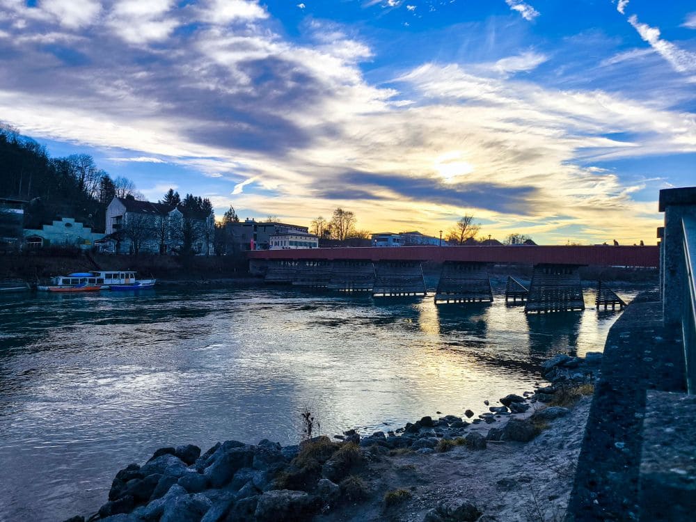 Bei Sonnenuntergang präsentiert sich die Flusslandschaft von Wasserburg mit einer Holzbrücke. Links ruht ein Boot, während sich Gebäude vor einem leuchtenden Himmel mit Wolken in Blau-, Weiß- und Goldtönen abheben. Im Vordergrund liegt das felsige Flussufer ruhig.