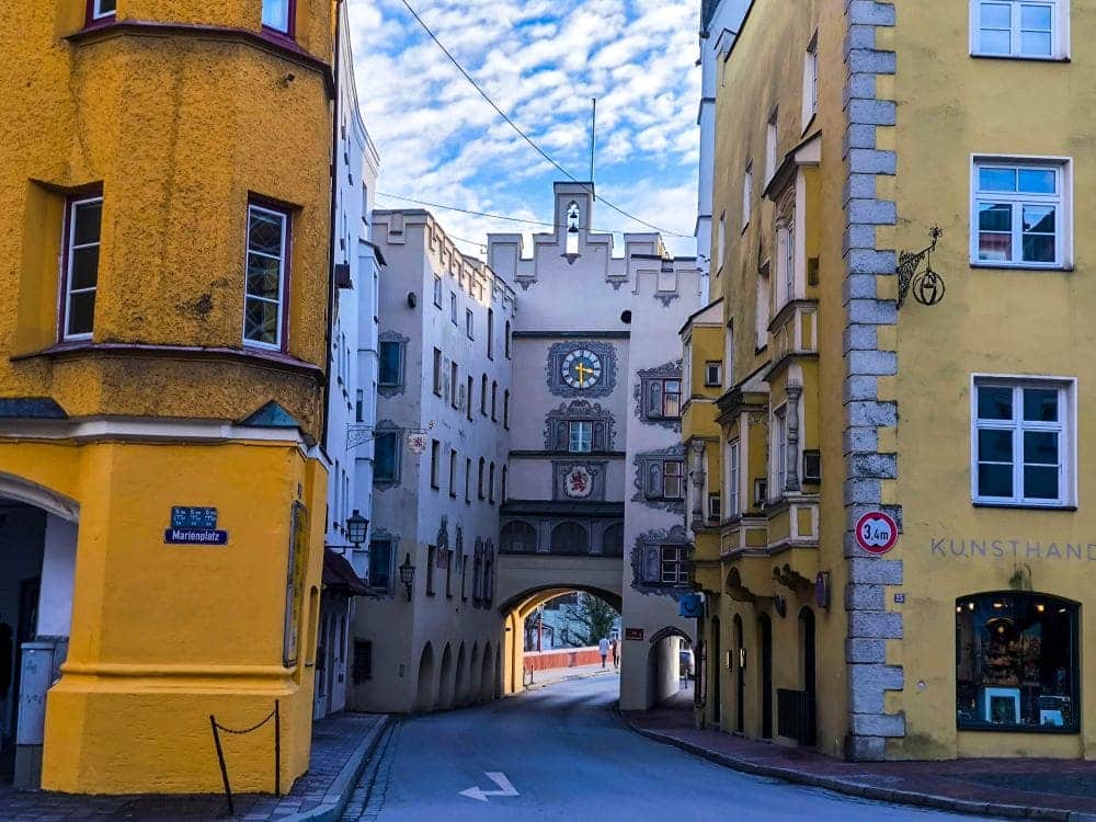 Eine schmale Straße in Wasserburg, gesäumt von farbenfrohen Gebäuden, führt zu einem kunstvollen Torbogen unter einem Glockenturm. Leuchtend gelbe und weiße Fassaden rahmen den Blick ein, während sich darüber ein teilweise bewölkter blauer Himmel erstreckt.