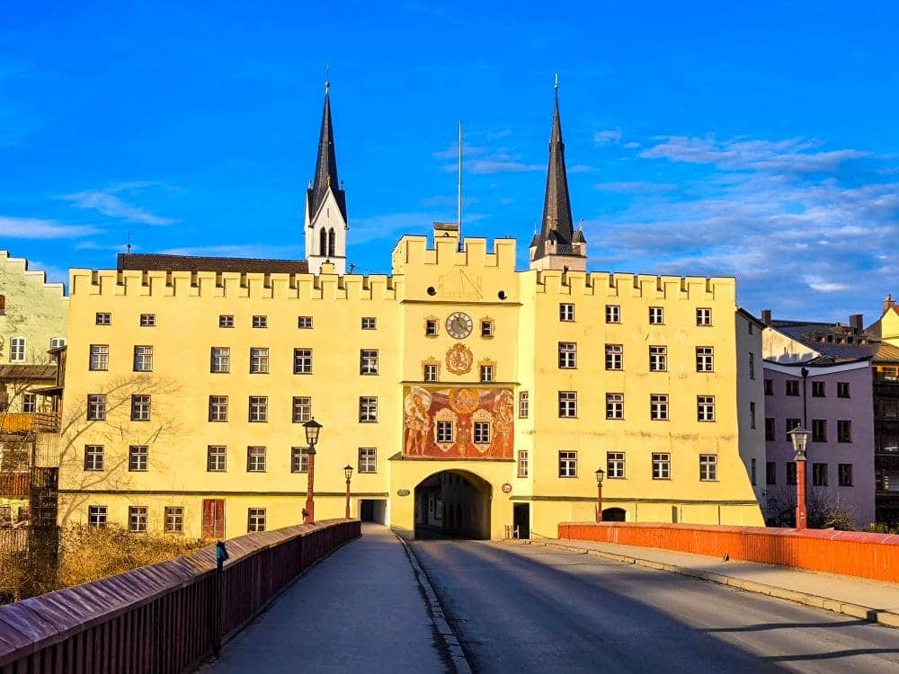 Unter strahlend blauem Himmel erhebt sich ein schlossähnliches Gebäude, das an eine Wasserburg erinnert und zwei hohe Türme besitzt. Zahlreiche Fenster und ein detailreiches Wappen in der Mitte sind durch eine Brücke verbunden, die in einen Torbogen mündet.