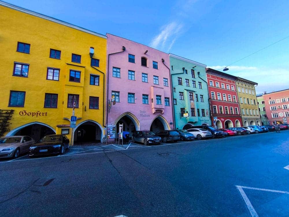 Eine Reihe farbenfroher Gebäude, die an das charmante Wasserburg erinnern, säumen die Straße. Davor parken Autos. Die Fassaden leuchten in Gelb-, Orange-, Rosa-, Blaugrün- und Rottönen unter einem strahlend blauen Himmel. Rechts in der weitgehend leeren Straße steht eine Straßenlaterne.