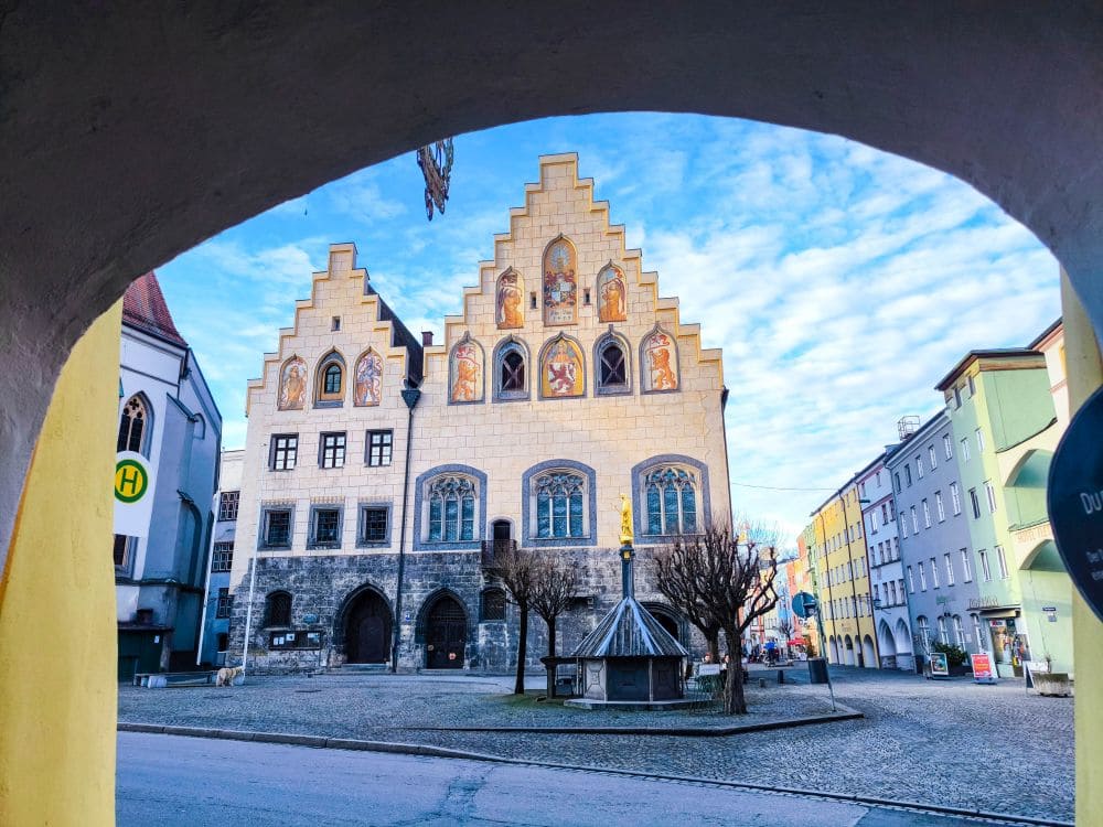 Ein historisches Gebäude mit heller Fassade und Stufengiebeln steht auf einem gepflasterten Platz, der an den alten Wasserburg-Stil erinnert und von einem Torbogen eingerahmt wird. Der Himmel ist teilweise bewölkt, blattlose Bäume umgeben das Gebäude. In der Nähe sind moderne Gebäude zu sehen.