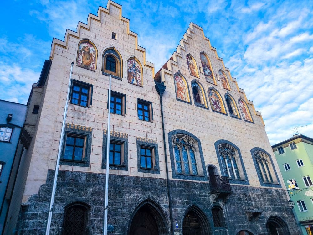 Eine historische Wasserburg mit einer Steinfassade, die aufwendige Muster und farbenfrohe religiöse Fresken auf Stufengiebeln aufweist, thront majestätisch. Der klare, mit vereinzelten Wolken durchsetzte Himmel unterstreicht die architektonischen Details, während moderne Gebäude durch die Landschaft ragen.