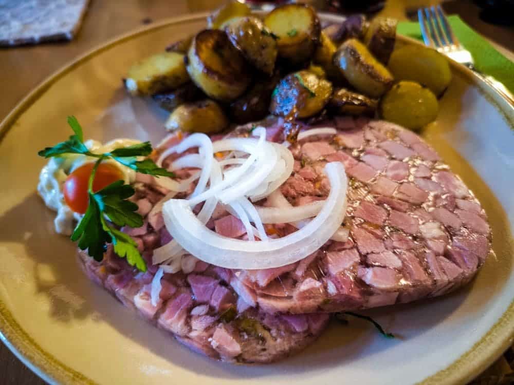 Ein Tellergericht mit zwei Scheiben Noahs Segel-Sülze mit Hackfleisch, garniert mit rohen Zwiebelringen, serviert mit Bratkartoffeln, einer Kirschtomate, Petersilie und einem Klecks Sahnesauce.