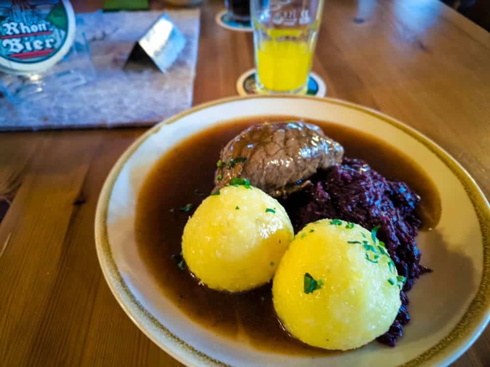 Ein Teller mit zwei Kartoffelknödeln, geschmortem Rindfleisch in brauner Soße und Rotkohl. Auf dem Holztisch steht ein Glas mit gelbem Getränk neben einem Untersetzer mit Rhön-Bier-Schriftzug und einer Noahs-Segel-Karte im Hintergrund.