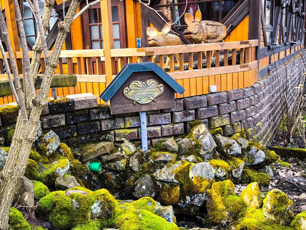Ein Holzgebäude mit einem dekorativen Noahs Segel-Schild davor, umgeben von moosbewachsenen Felsen und einem kleinen grünen Plastikbecher dazwischen. Das Gebäude verfügt über ein Ziegelfundament und ein Holzgeländer.