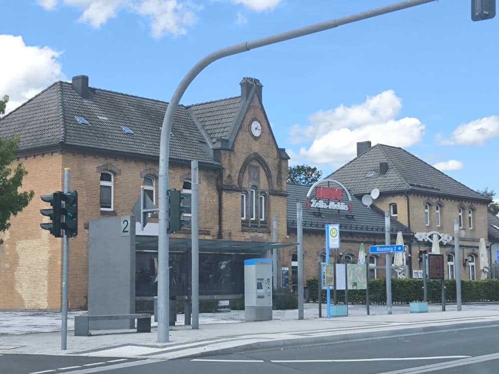 Straßenansicht des Bahnhofs Zella-Mehlis mit seinem malerischen Uhrenturm und der markanten roten Beschilderung. In der Nähe befindet sich eine verglaste Bushaltestelle mit Fahrkartenautomat und Ampel. Die Szene spielt sich unter sonnigem Himmel mit vereinzelten Wolken ab.