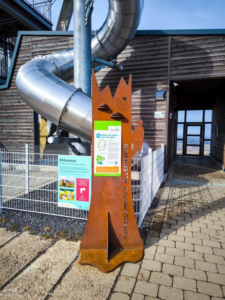 Eine rostfarbene Metallstatue in Form einer lächelnden Figur steht neben einem Informationsschild in der Nähe des modernen Holzgebäudes von Noahs Segel, an dessen Außenseite sich eine große silberne Röhrenrutsche befindet. Der Boden ist gepflastert und eingezäunte Bereiche sind sichtbar.