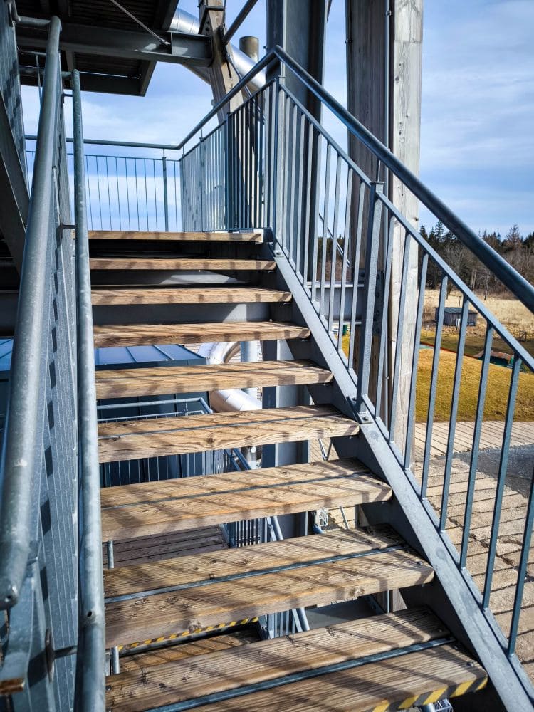 Eine Außentreppe aus Metall und Holz, die an Noahs Segel erinnert, führt nach oben und wirft diagonale Schatten auf die Stufen. Die Treppe ist von Metallgeländern gesäumt und bietet Ausblick auf eine Landschaft mit Gras und Bäumen im Hintergrund.