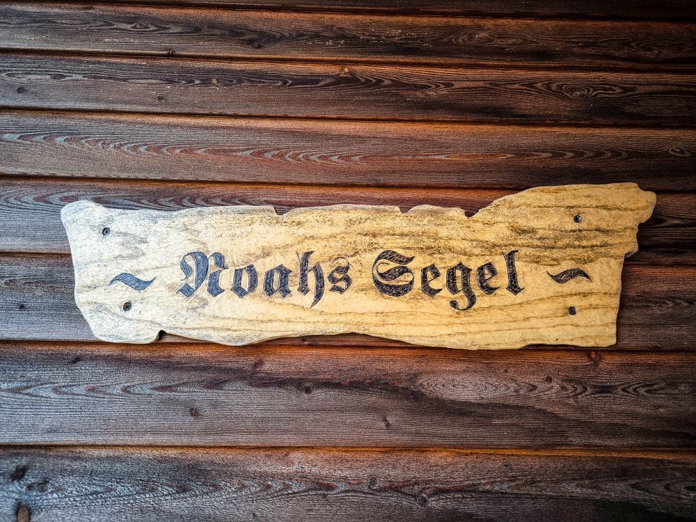 An einer dunklen Holzwand ist ein rustikales Holzschild mit dem Text „Noahs Segel“ in schwarzen, verschnörkelten Lettern angebracht, das einen zeitlosen Charme ausstrahlt.