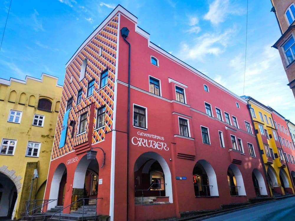 Die farbenfrohe Wasserburg besticht durch geometrische Muster und Bogenfenster vor einer markanten roten Fassade mit weiß-orange gemusterten Bereichen. Mehrere Gedenktafeln schmücken die Wände, während der klare Himmel mit einigen zarten Wölkchen aufwartet.