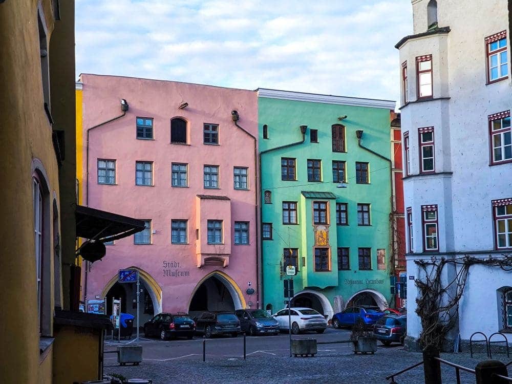 Bunte Gebäude, die an eine malerische Wasserburg erinnern, säumen die gepflasterte Straße. Ihre rosa und grünen Fassaden und die gewölbten Eingänge zieren die Straße. Davor parken Autos, während der leicht bewölkte Himmel den historischen Charme noch verstärkt. Zahlreiche Fenster und dekorative Elemente runden das Bild ab.