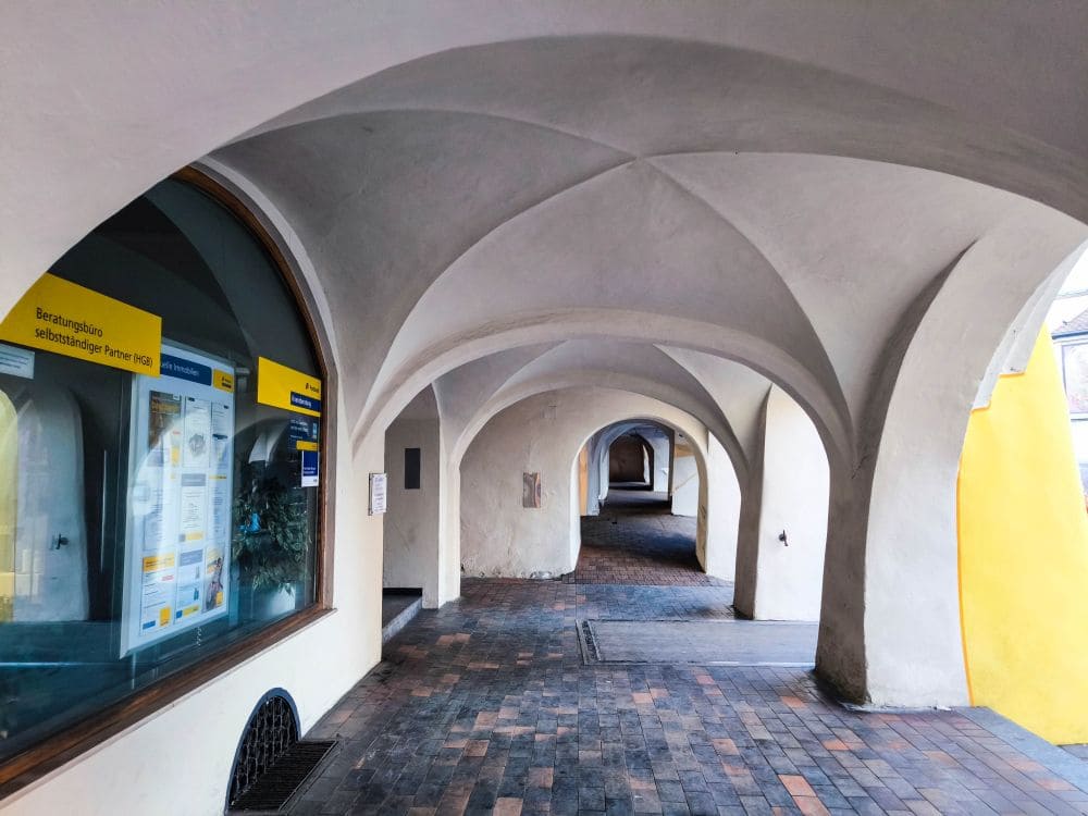 Ein überdachter, gewölbter Gang mit gefliestem Boden und cremefarbenen Wänden erinnert an die charmante Architektur von Wasserburg. Ein großes Fenster links zeigt ein gelbes Schild mit Text. Sonnenlicht wirft Schatten auf den Weg und führt zu einem einladenden Torbogen in der Ferne.