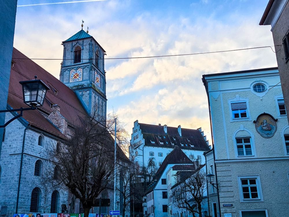 Eine malerische Straßenansicht in Wasserburg zeigt eine alte Steinkirche mit Glockenturm unter blauem Himmel mit Wolken. Die angrenzenden Gebäude bestechen durch ihre klassische Architektur, und kahle Bäume säumen die Straße. Eine alte Straßenlaterne ziert den Vordergrund und unterstreicht den zeitlosen Charme der Stadt.