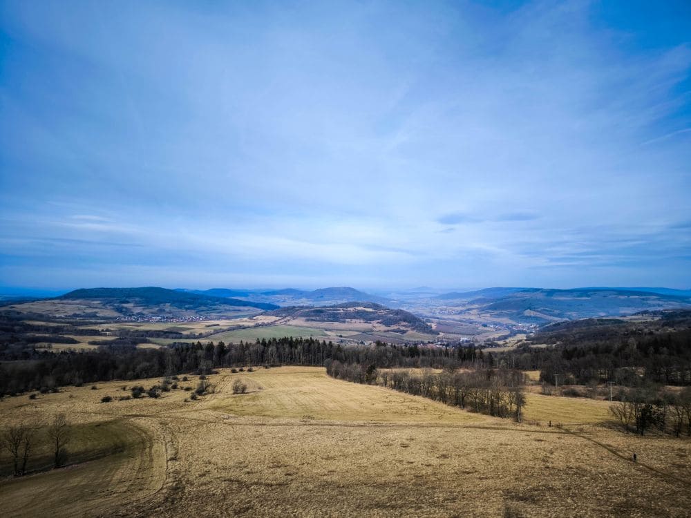 Eine weite Landschaft mit sanften Hügeln und offenen Feldern unter blauem Himmel mit zarten Wolken. Vereinzelte Bäume und Waldstücke prägen die Landschaft, und in der Ferne erhebt sich Noahs Segel unter fernen Bergen.