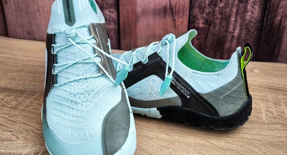 Ein Paar hellblaue Sportschuhe mit schwarz-grünem Absatz, gefertigt für ein Barfußgefühl mit minimaler Fußunterstützung. Sie bestehen aus strukturiertem Strickstoff mit Schnürsenkeln vor einem rustikalen Holzhintergrund.