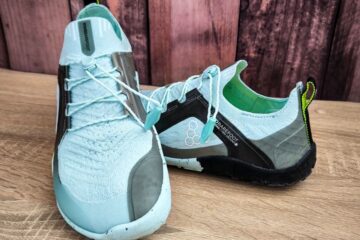 Ein Paar hellblaue Sportschuhe mit schwarz-grünem Absatz, gefertigt für ein Barfußgefühl mit minimaler Fußunterstützung. Sie bestehen aus strukturiertem Strickstoff mit Schnürsenkeln vor einem rustikalen Holzhintergrund.