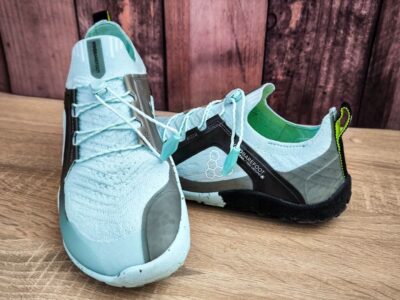 Ein Paar hellblaue Sportschuhe mit schwarz-grünem Absatz, gefertigt für ein Barfußgefühl mit minimaler Fußunterstützung. Sie bestehen aus strukturiertem Strickstoff mit Schnürsenkeln vor einem rustikalen Holzhintergrund.
