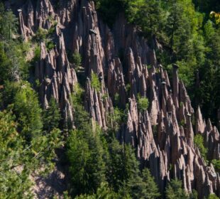 Eine Naturlandschaft mit den hoch aufragenden Erdpyramiden, deren hohe, schmale, turmähnliche Felsformationen hervorstechen. Diese markanten Strukturen erheben sich inmitten eines dichten, grünen Waldes und bilden einen faszinierenden Kontrast zwischen den rauen Felsstrukturen und dem üppigen Laub.