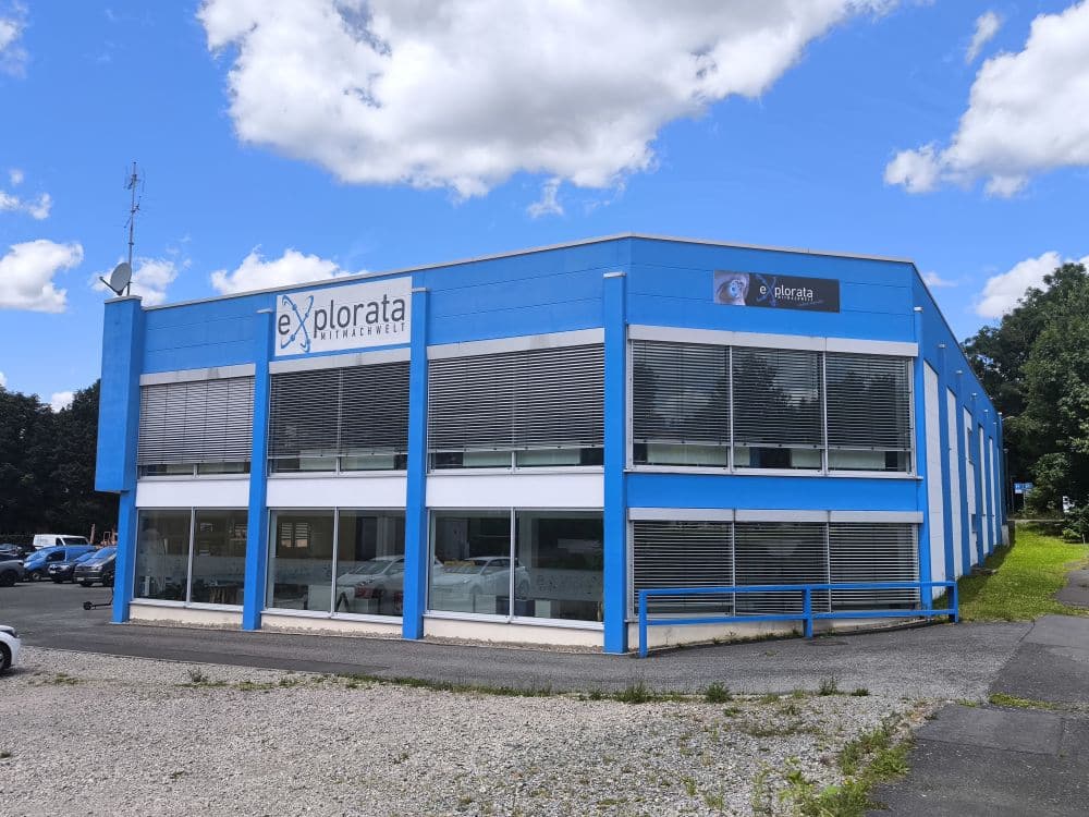 Ein zweistöckiges Gebäude in Zella-Mehlis besticht durch seine blau-weiße Fassade unter leicht bewölktem Himmel. Große Fenster schmücken beide Etagen, und die „explorata“-Beschilderung ziert die Fassade. Links parken mehrere Autos, was den Charme der Szenerie unterstreicht.