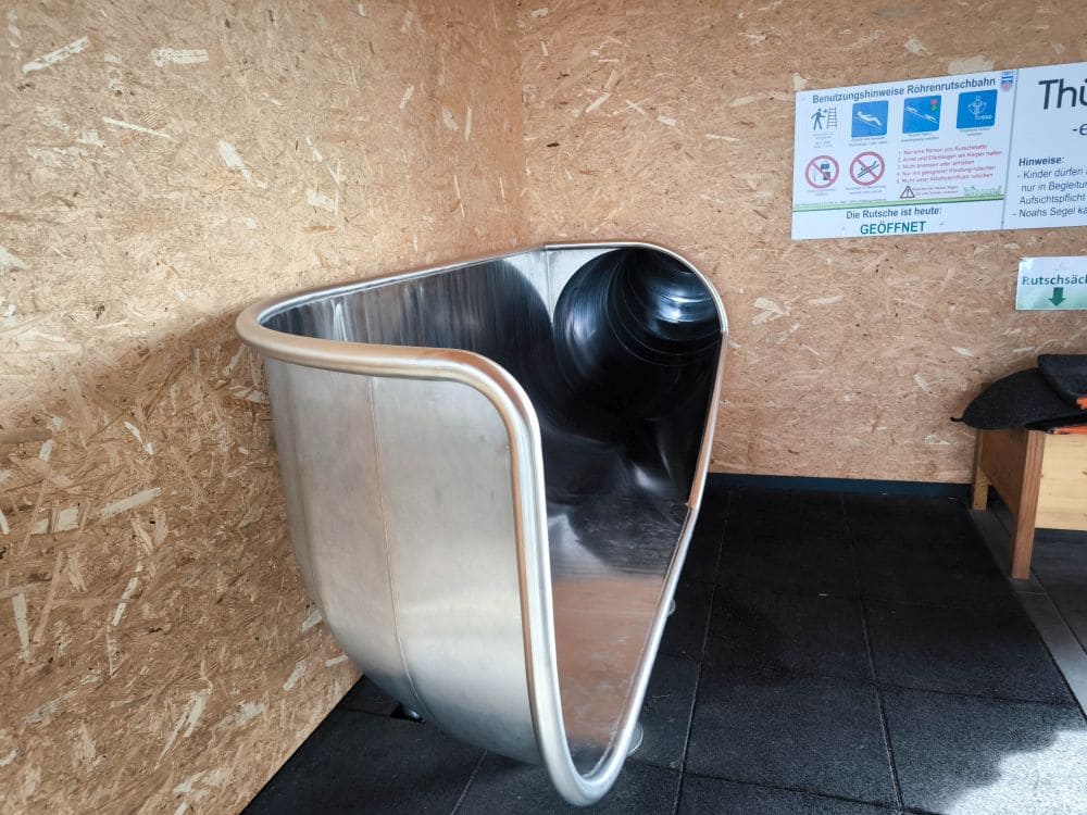 Ein glänzender Metall-Röhrenrutschen-Eingang im Innenbereich, vor Holzwänden mit darüber angebrachten Schildern, verleiht dem Ganzen eine verspielte Note, die an Noahs Segel erinnert. Die Rutsche hat eine abgerundete Kante und einen schwarzen Gummiboden davor.