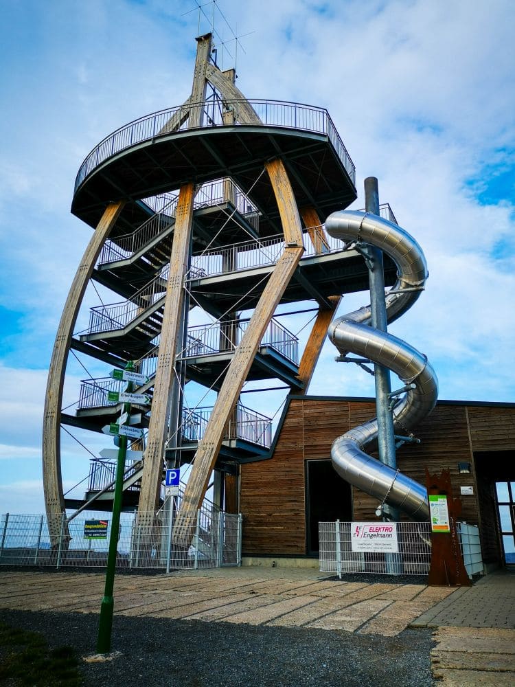 Noahs Segel, ein hoher Aussichtsturm aus Holz und Metall mit einer silbernen gewundenen Rutsche, mehreren Treppen und einem umzäunten Sockel, steht neben einem Holzgebäude unter einem teilweise bewölkten Himmel.