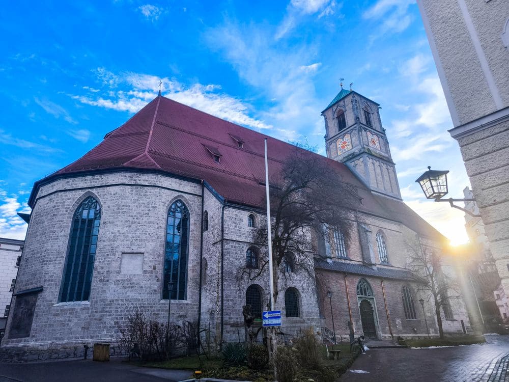 Eine große Steinkirche mit rotem Ziegeldach und Glockenturm steht unter dem strahlend blauen Himmel von Wasserburg. Die Sonne lugt hervor und wirft einen warmen Schein, während blattlose Bäume und eine Straßenlaterne die Szenerie einrahmen und eine friedliche Morgenstimmung schaffen.