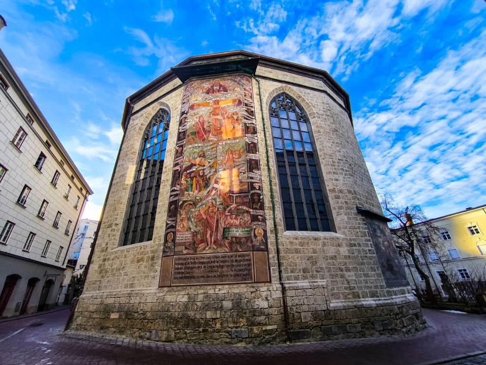 Ein historisches Steingebäude, das an eine Wasserburg erinnert, besticht durch hohe Bogenfenster und ein lebendiges Wandgemälde mit religiösen Szenen. Der strahlend blaue Himmel mit vereinzelten Wolken unterstreicht seine Pracht. Umgeben von Gebäuden umrundet ein Kopfsteinpflasterweg das Gebäude.