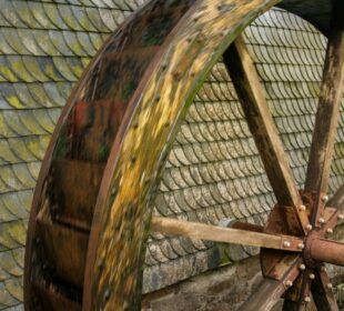 Nahaufnahme des Wasserrads der Oberwallbachsmühle. Seine großen Holzschaufeln sind teilweise mit Moos bedeckt. Das Rad steht vor einem Hintergrund aus alten, moosbedeckten Steinschindeln und unterstreicht den historischen Charme dieses bemerkenswerten Bauwerks.