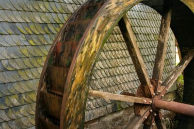 Nahaufnahme des Wasserrads der Oberwallbachsmühle. Seine großen Holzschaufeln sind teilweise mit Moos bedeckt. Das Rad steht vor einem Hintergrund aus alten, moosbedeckten Steinschindeln und unterstreicht den historischen Charme dieses bemerkenswerten Bauwerks.