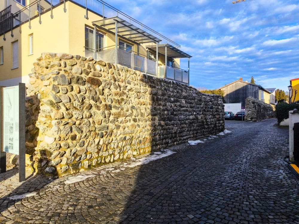 Eine Steinmauer säumt eine Kopfsteinpflasterstraße im charmanten Wohnviertel Wasserburg. Ein Gebäude mit Balkonen ziert die linke Seite, während darüber ein leicht bewölkter Himmel schwebt. Die Sonne wirft verspielte Schatten, die die reiche Textur der Steine hervorheben und den malerischen Reiz der Szenerie verstärken.