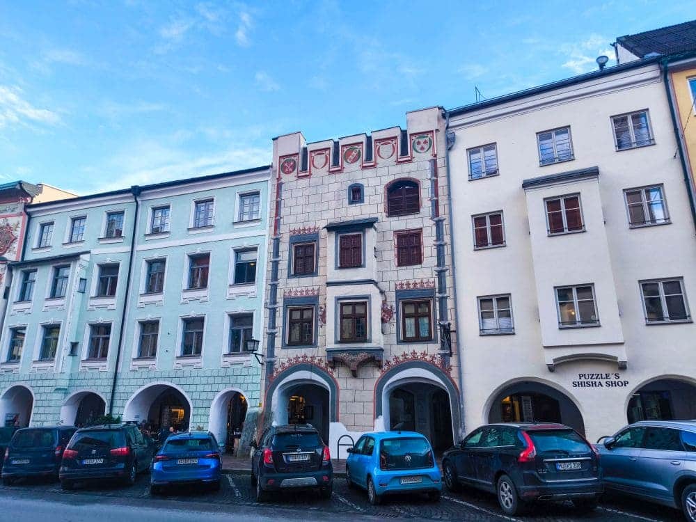 Straßenansicht einer Reihe historischer europäischer Gebäude in Wasserburg mit gewölbten Eingängen und kunstvollen Verzierungen. Vor dem Gebäude parken Autos, und verschiedene Geschäfte wie der Puzzles Shisha Shop befinden sich im Inneren. Über dem Gebäude erstreckt sich ein strahlend blauer Himmel.