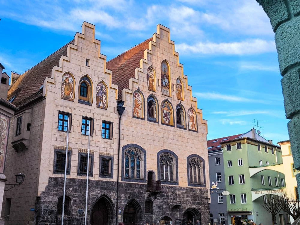 Die historische Wasserburg besticht durch ihr Giebeldach und die dekorativen Fresken an der Fassade. Ihre Bogenfenster und das kunstvolle Mauerwerk bilden einen Kontrast zu den modernen Gebäuden in der Nähe, die alle unter einem blauen Himmel mit zarten Wolken stehen.