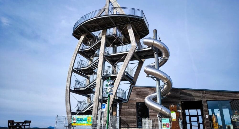 Ein moderner Aussichtsturm, bekannt als Noahs Segel, mit Wendeltreppe und großer Spiralröhrenrutsche steht neben einem Holzgebäude unter blauem Himmel. Vor diesem markanten Bauwerk stehen ein Picknicktisch und mehrere Schilder.