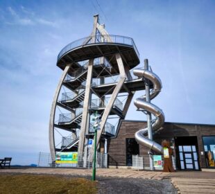 Ein moderner Aussichtsturm, bekannt als Noahs Segel, mit Wendeltreppe und großer Spiralröhrenrutsche steht neben einem Holzgebäude unter blauem Himmel. Vor diesem markanten Bauwerk stehen ein Picknicktisch und mehrere Schilder.