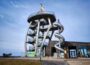 Ein moderner Aussichtsturm, bekannt als Noahs Segel, mit Wendeltreppe und großer Spiralröhrenrutsche steht neben einem Holzgebäude unter blauem Himmel. Vor diesem markanten Bauwerk stehen ein Picknicktisch und mehrere Schilder.