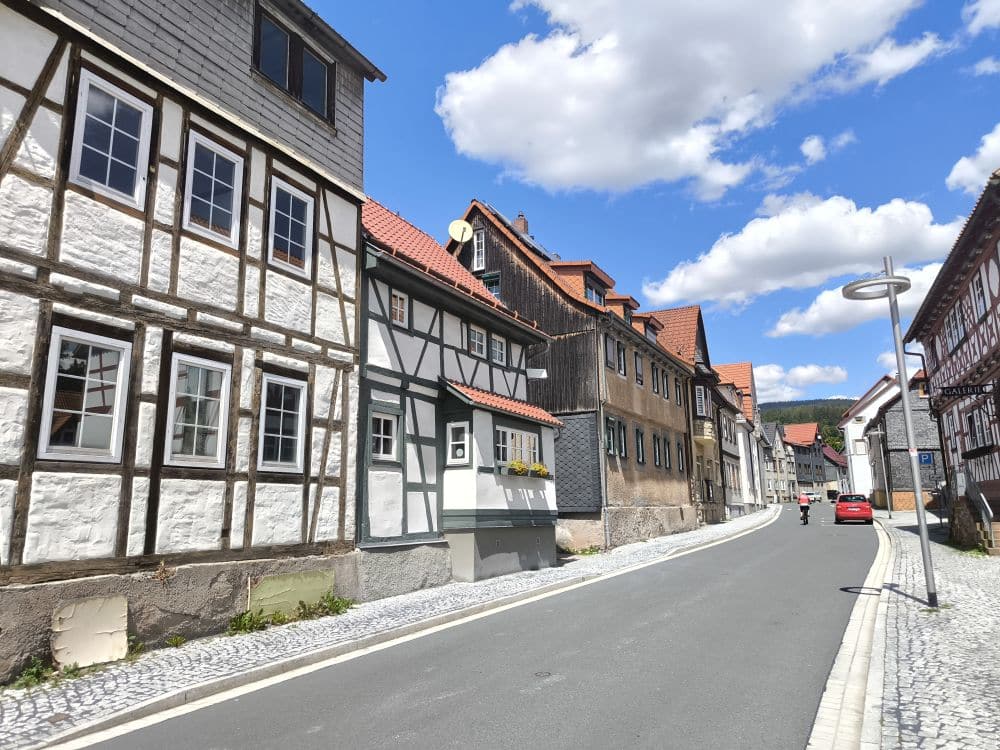 Eine malerische Straße in Zella-Mehlis ist gesäumt von traditionellen Fachwerkhäusern unter einem strahlend blauen Himmel mit flauschigen Wolken. Ein Mensch schlendert die Straße entlang, rechts steht ein Laternenpfahl. Die Gebäude bestechen durch dekoratives Holz und weißen Putz und versprühen zeitlosen Charme.