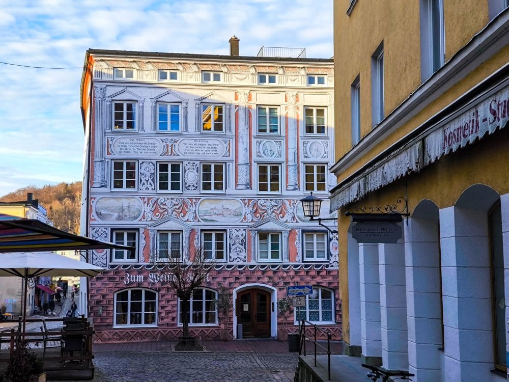 Ein farbenfrohes Gebäude mit aufwendigen Malereien, darunter Muster und Säulen, die an eine klassische Wasserburg erinnern. Es liegt an einer Kopfsteinpflasterstraße und ist teilweise im Schatten, während rechts ein weiteres gelbes Gebäude mit einem gewölbten Eingang steht.