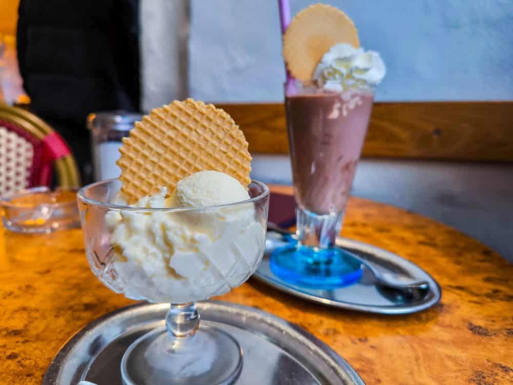 Eine Kugel Vanilleeis mit Waffel in einer Glasschale auf einem Tablett, dazu ein Schokoladenmilchshake mit Schlagsahne und Waffel, elegant serviert auf einem blauen Teller mit Löffel. Diese reizvolle Szene findet man in charmanten Cafés in ganz Wasserburg.