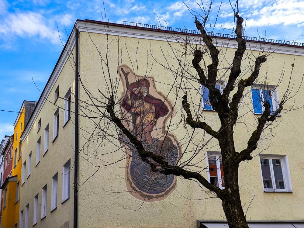 Ein kahler Baum steht vor einem gelben Gebäude in Wasserburg. Das Wandgemälde zeigt eine gekleidete Gestalt mit einem Stab in der Hand. Der Himmel ist strahlend blau mit leichten Wolken, und mehrere Fenster durchbrechen die Fassade des Gebäudes.