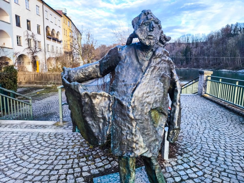 Eine Bronzestatue in Wasserburg zeigt eine Person mit strukturiertem, abstraktem Gesicht, die einen Mantel trägt und mit ausgestrecktem Arm gestikuliert. Sie steht auf einem Kopfsteinpflasterweg in der Nähe eines Geländers, eingerahmt von Gebäuden und Bäumen unter einem teilweise bewölkten Himmel.