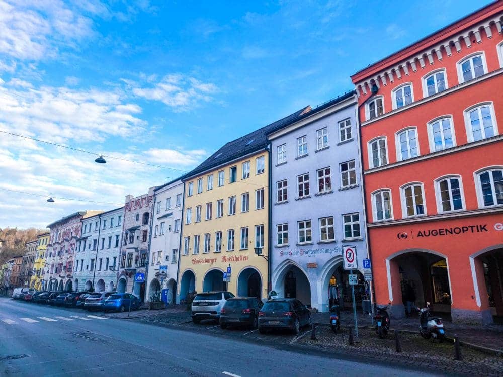 Straßenansicht mit farbenfrohen Gebäuden im europäischen Stil und gewölbten Eingängen, die an die charmante Wasserburger Architektur erinnern. Autos und Motorräder säumen den Bürgersteig, während ein klarer blauer Himmel mit einigen Wolken darüber schwebt.