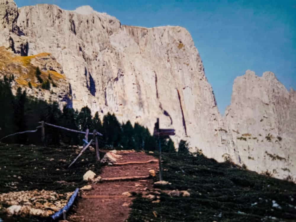 Ein felsiger Bergpfad mit Holzzaun und Wegweiser führt zu den hoch aufragenden, sonnenbeschienenen Felsen des Schlern. Kiefern säumen den Weg, und der Himmel darüber ist klar und blau – perfekt für eine unvergessliche Wanderung voller Abenteuer.