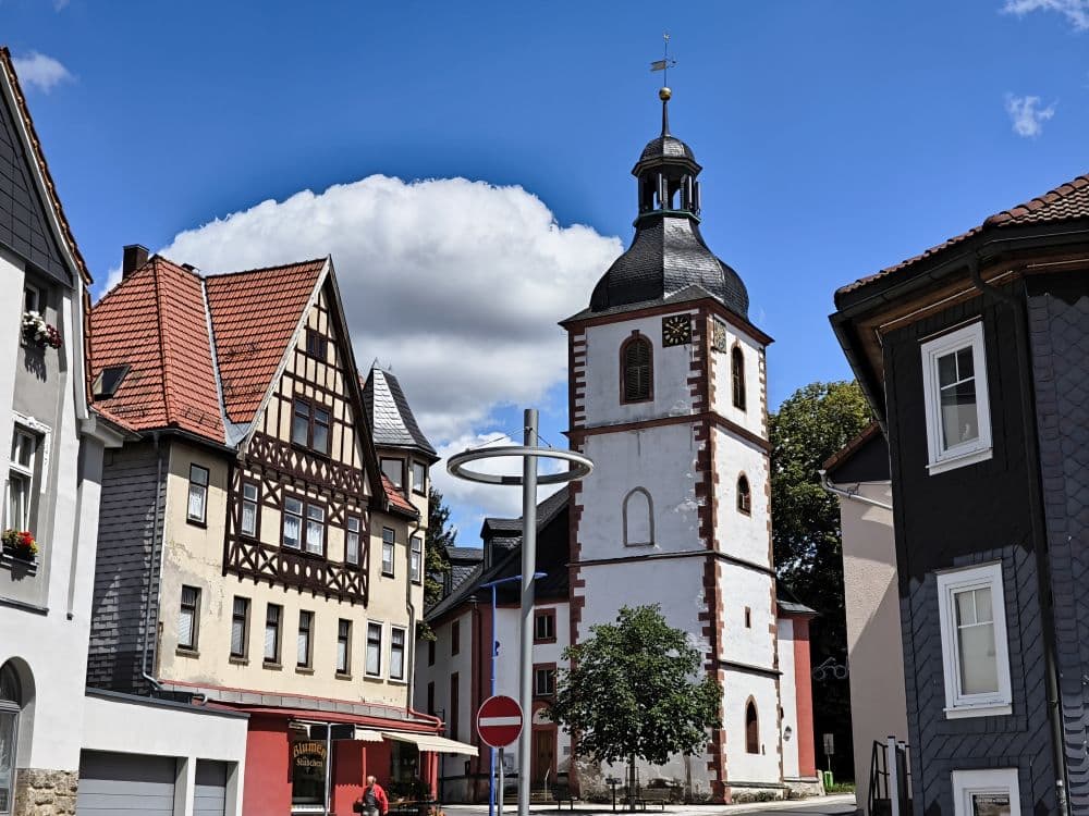 Eine charmante Straße in Zella-Mehlis besticht durch traditionelle Fachwerkhäuser und eine weiße Kirche mit Kirchturm. Links ist ein Schild mit der Aufschrift „Einfahrt verboten“ zu sehen. Der Himmel darüber ist strahlend blau und mit großen, flauschigen Wolken geschmückt.