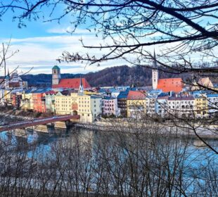 Ein malerischer Blick auf die farbenfrohe Flussstadt Wasserburg am Inn, deren charmante Gebäude von ihrer Geschichte zeugen. Eine Brücke überspannt elegant das Wasser und führt Sie an einer Kirche mit hoch aufragendem Kirchturm vorbei. Kahle Äste rahmen diese malerische Szenerie vor einer bewaldeten Hügelkulisse ein.