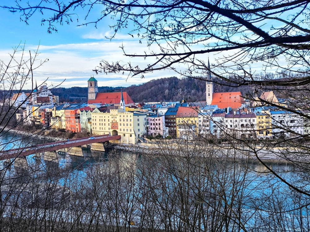 Ein malerischer Blick auf die farbenfrohe Flussstadt Wasserburg am Inn, deren charmante Gebäude von ihrer Geschichte zeugen. Eine Brücke überspannt elegant das Wasser und führt Sie an einer Kirche mit hoch aufragendem Kirchturm vorbei. Kahle Äste rahmen diese malerische Szenerie vor einer bewaldeten Hügelkulisse ein.
