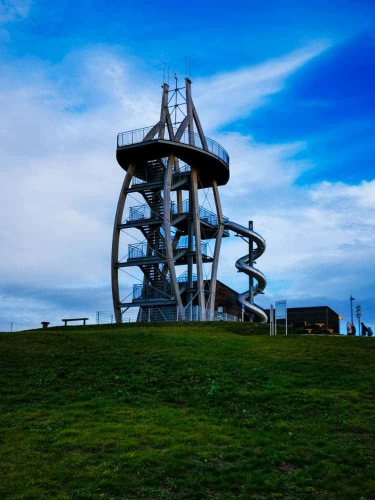 Ein moderner Aussichtsturm aus Metall, bekannt als Noahs Segel, mit einer außenliegenden Spiralrutsche steht auf einem grasbewachsenen Hügel unter einem blauen Himmel mit vereinzelten Wolken. In der Nähe des markanten Bauwerks sind mehrere Personen und Bänke zu sehen.