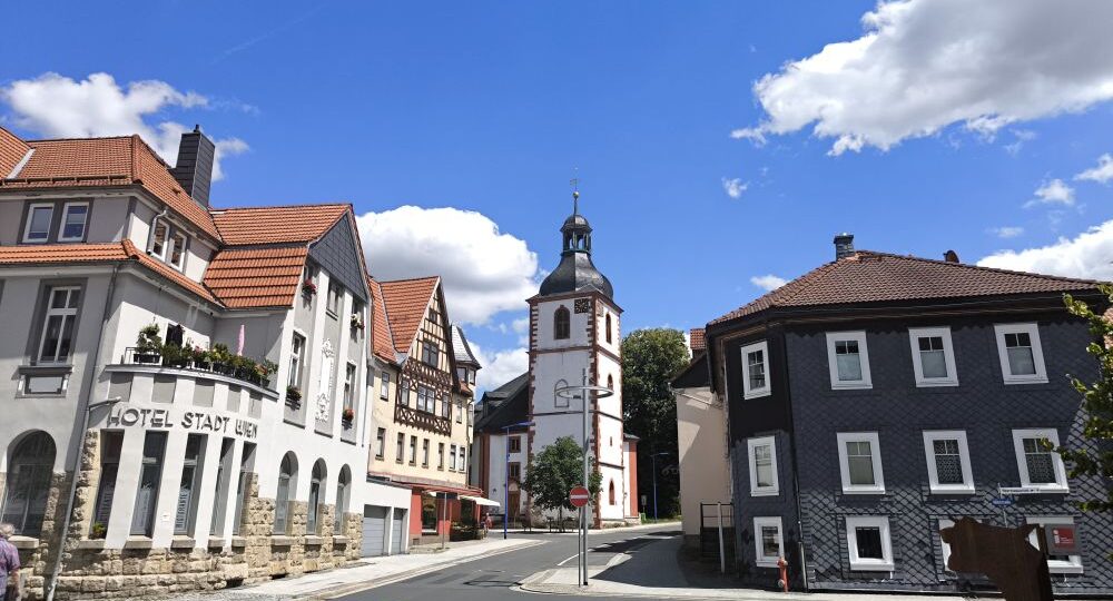 Im Herzen von Zella-Mehlis gelegen, bezaubert diese malerische Kleinstadtstraße mit ihrer traditionellen europäischen Architektur und dem historischen Kirchturm in der Mitte. Die malerischen Gebäude bestechen durch ihre roten Ziegeldächer vor dem Hintergrund eines blauen Himmels mit zarten, vereinzelten Wolken.