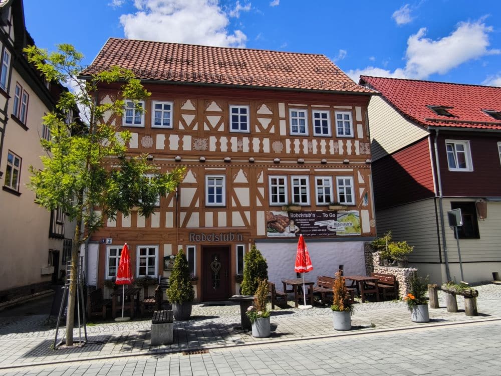 Ein charmantes, traditionelles Fachwerkhaus in Zella-Mehlis mit rotem Dach thront unter einem blauen, wolkenverhangenen Himmel. Zwei Tische mit roten Sonnenschirmen zieren die Kopfsteinpflasterstraße, gesäumt von Topfpflanzen. Die Fassade des Gebäudes besticht durch aufwendige Holzarbeiten.