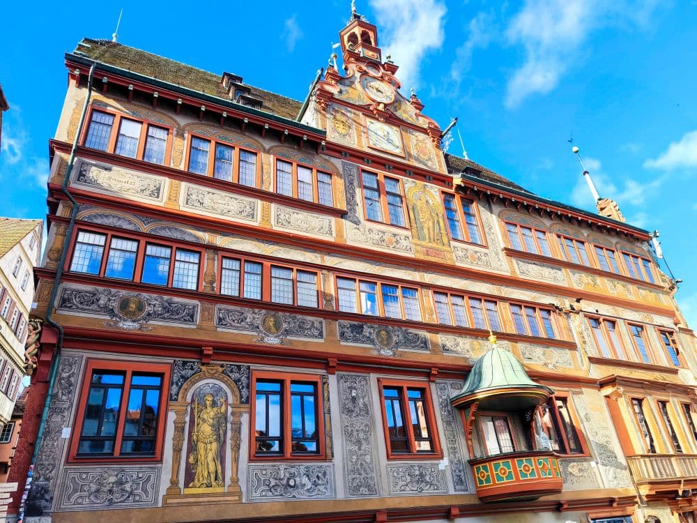 Reich verziertes historisches Gebäude in Tübingen mit detaillierten Wandmalereien, Statuen und rot gerahmten Fenstern unter blauem Himmel; verfügt über einen kleinen Balkon und eine große Uhr über dem Eingang.
